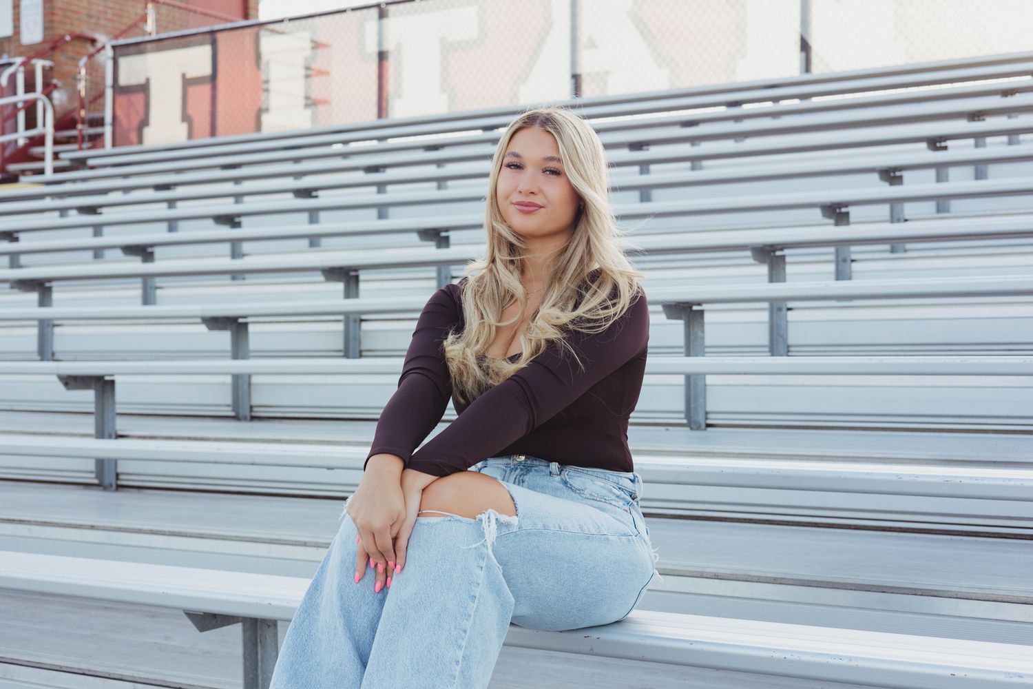 Blonde teenage girl in black top and jeans sits on bleachers for high school senior portrait photos.