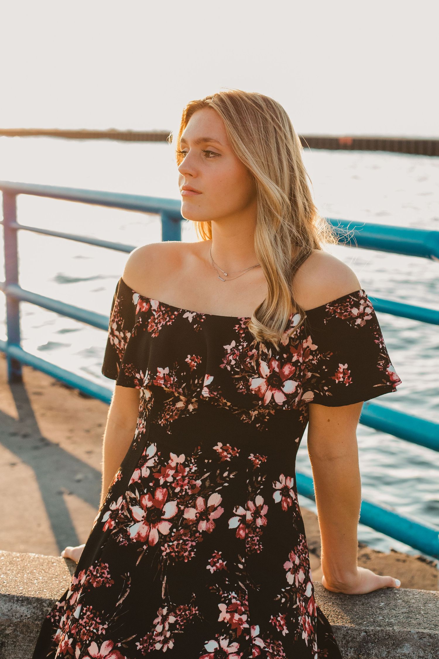 Young blonde woman in off-shoulder floral dress standing by waterfront railing at sunset.