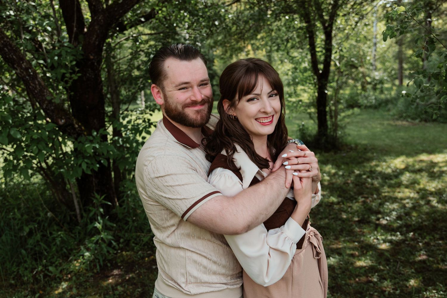 Happy couple embracing outdoors in lush green park, smiling woman with bangs and man with beard in casual attire.
