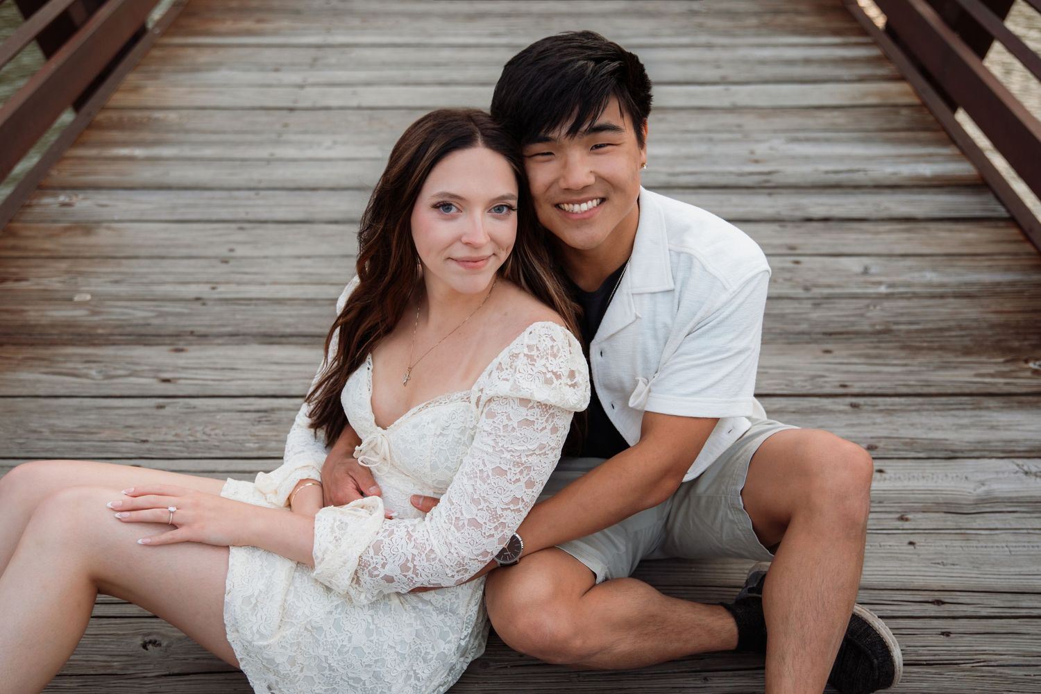 Happy couple sitting together on a wooden bridge, smiling warmly during their engagement photo session.
