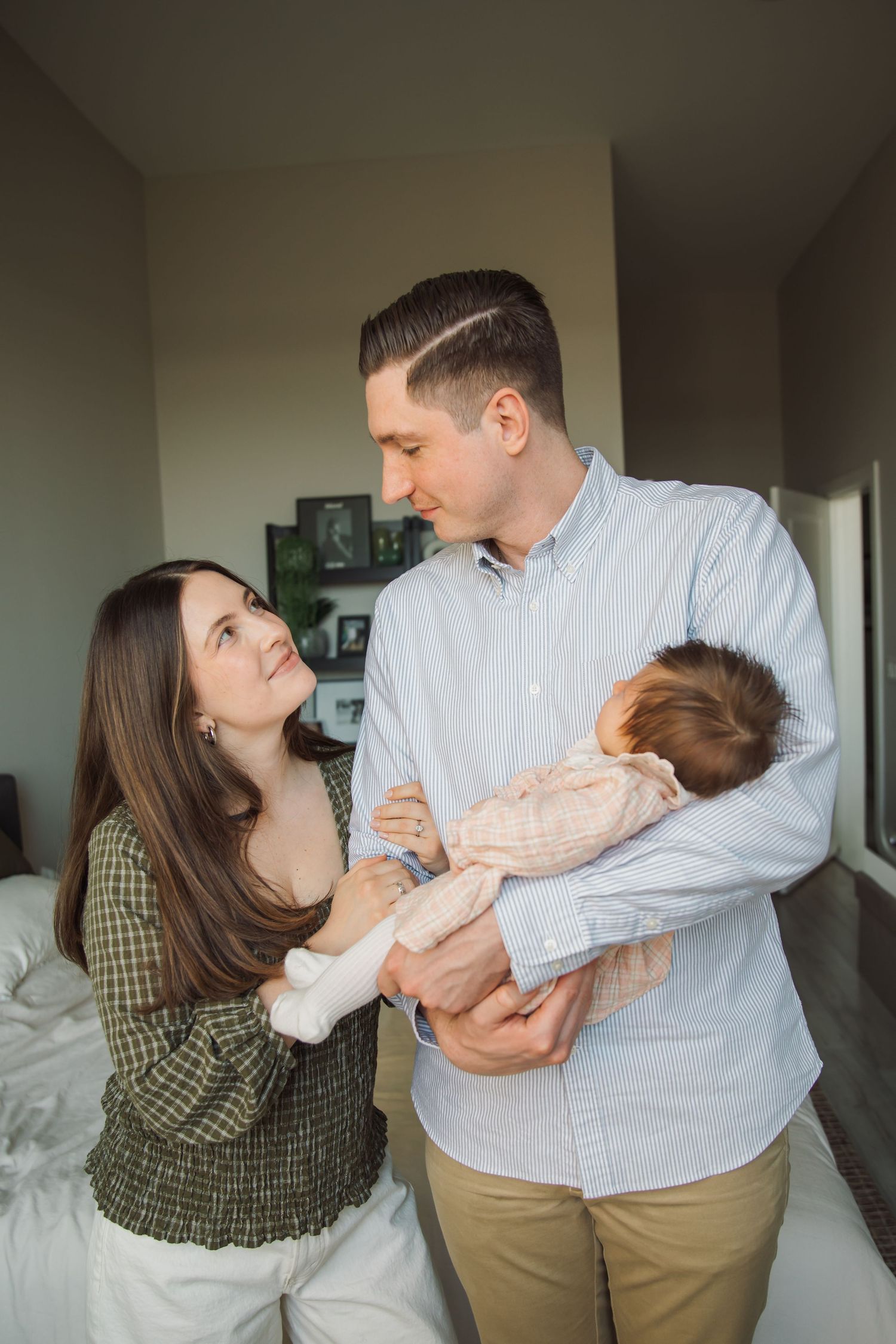 Loving parents holding newborn baby while gazing at each other in bedroom.