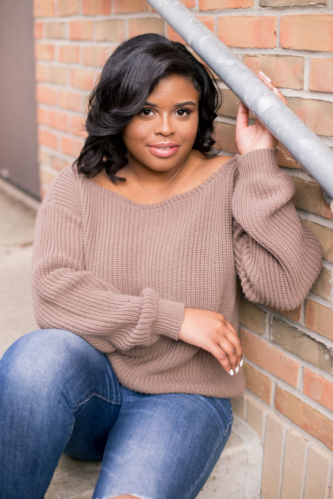 Young woman in brown knit sweater and jeans sits against a brick wall, holding a metal railing.