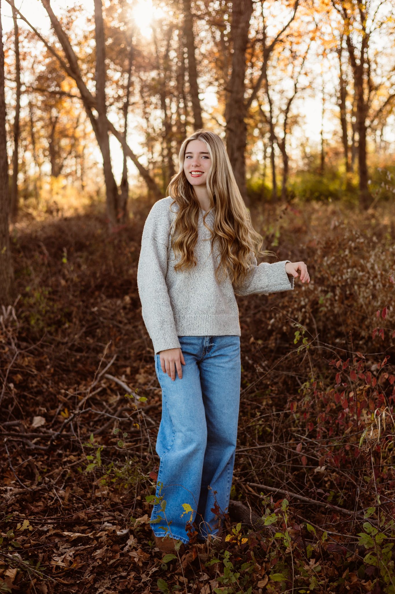 Teen girl in cream sweater and wide-leg jeans poses in autumn forest with golden sunlight filtering through trees.