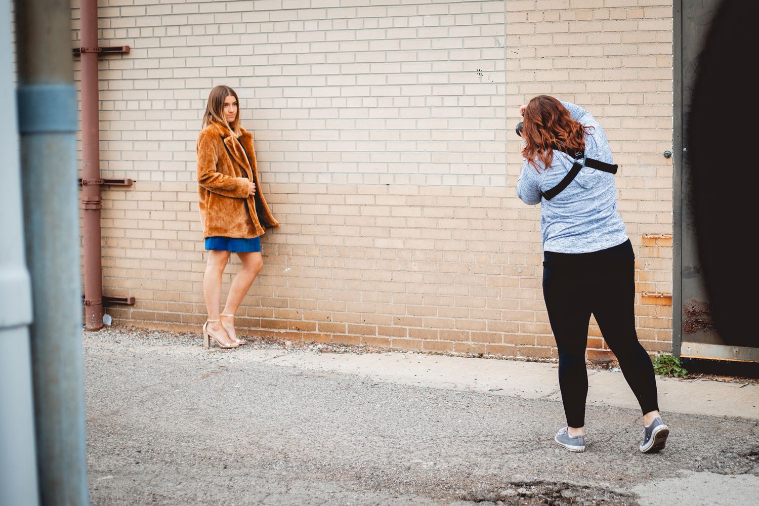 Photographer taking photos of a woman in an orange jacket and denim skirt posed against a brick wall outdoors.