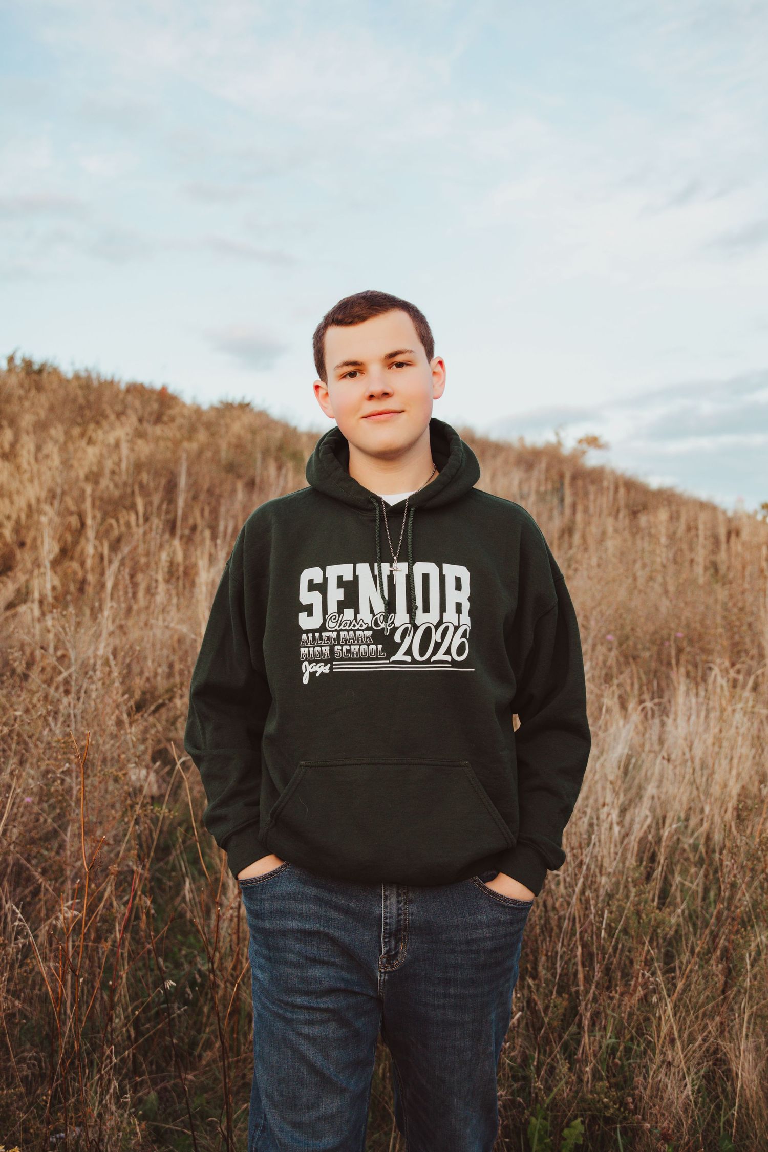 A teenage boy in a black Senior 2026 hoodie and jeans stands in a golden field under a cloudy sky.