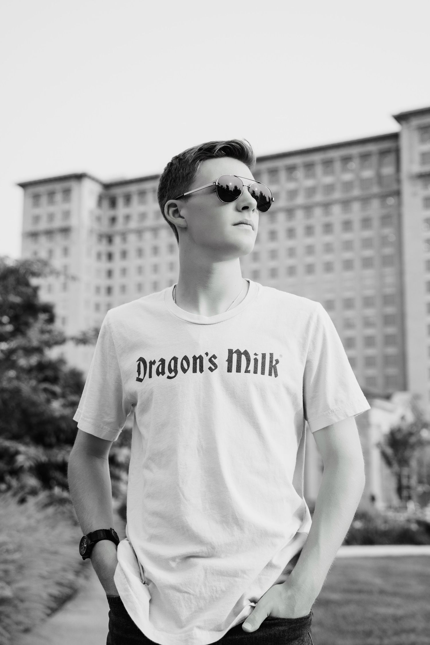 Young man in Dragon's Milk t-shirt wearing sunglasses standing outdoors near large building in black and white.
