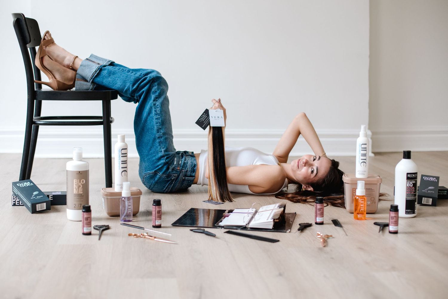 Woman lying on floor surrounded by hair care and beauty products with a chair nearby.