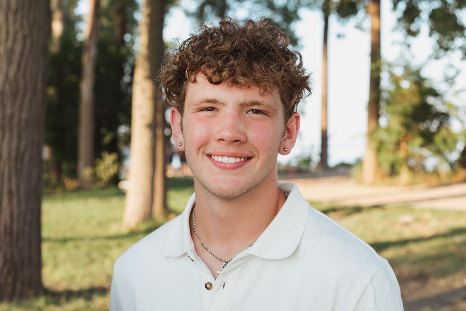 Smiling young man with curly hair wearing a white polo shirt outdoors among trees.