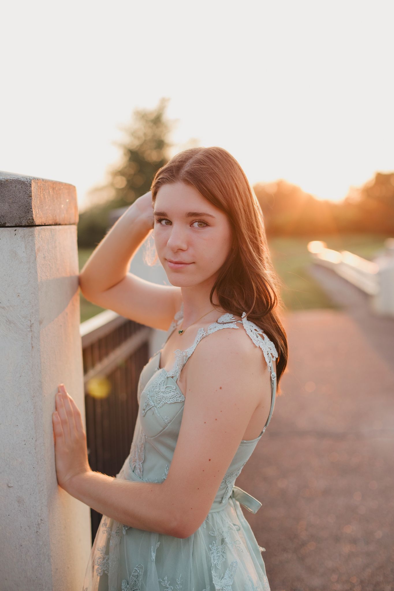 Young woman in mint green dress poses by concrete pillar during golden hour sunset on a bridge.