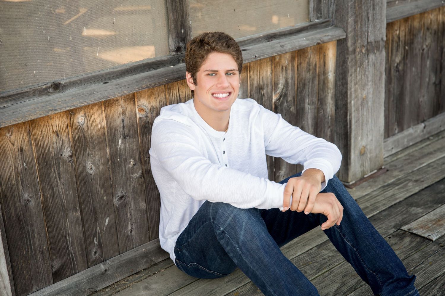 Young man in white henley and jeans smiling while sitting on wooden steps outdoors.
