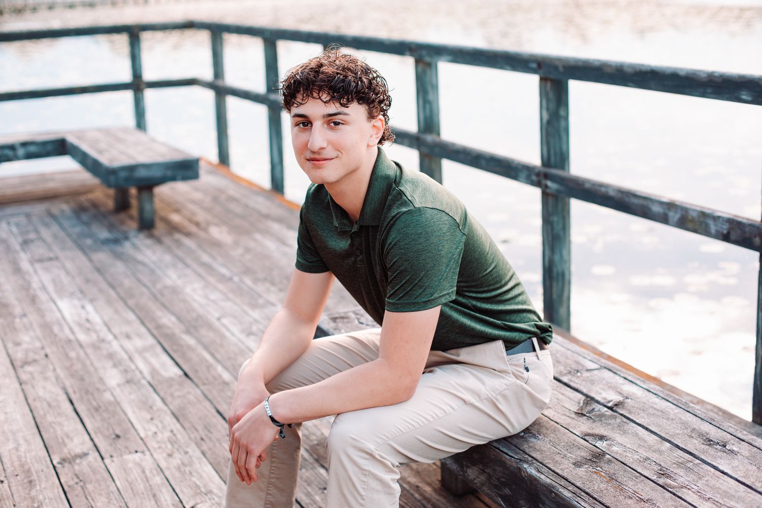 Young man in green polo shirt sitting on wooden dock by water, posing for senior portrait photo.