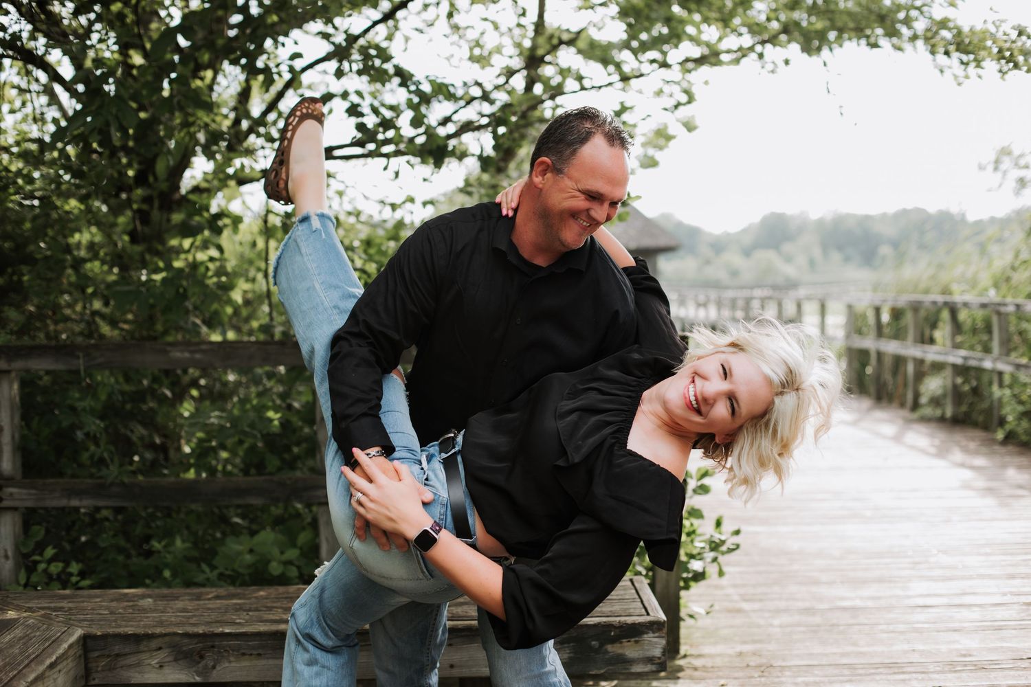 A playful couple laugh as the man dips the woman dramatically on a wooden bridge surrounded by trees.