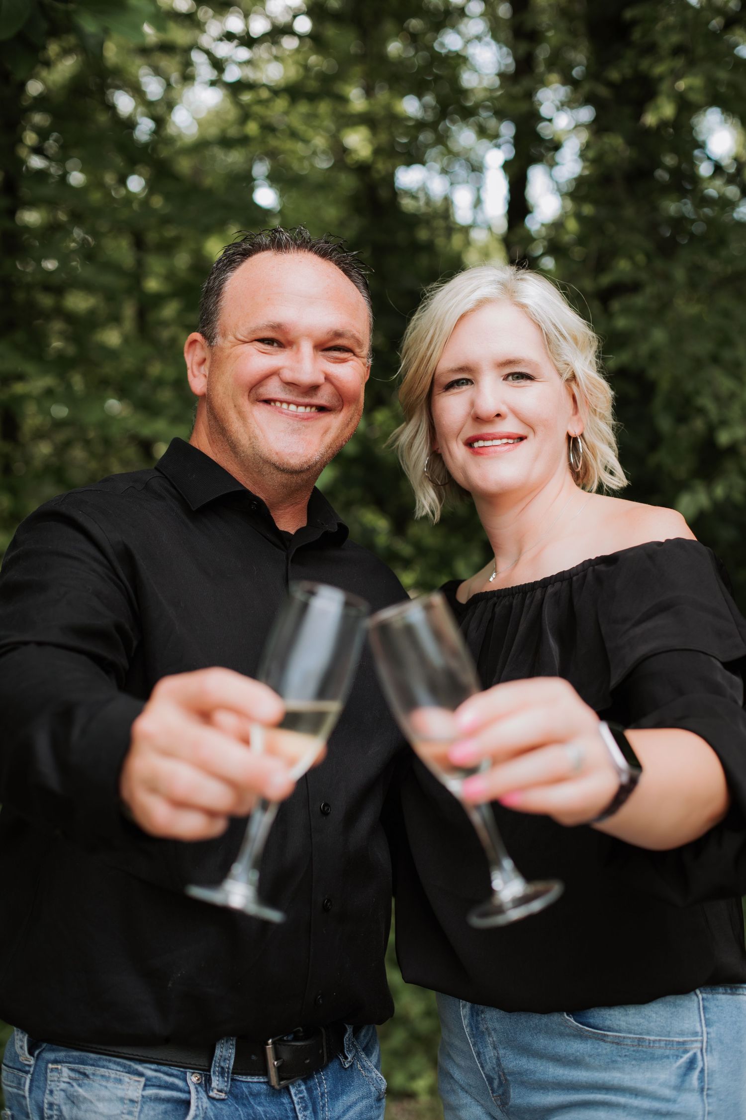 A happy couple smiling and toasting champagne glasses toward the camera in a lush green outdoor setting.