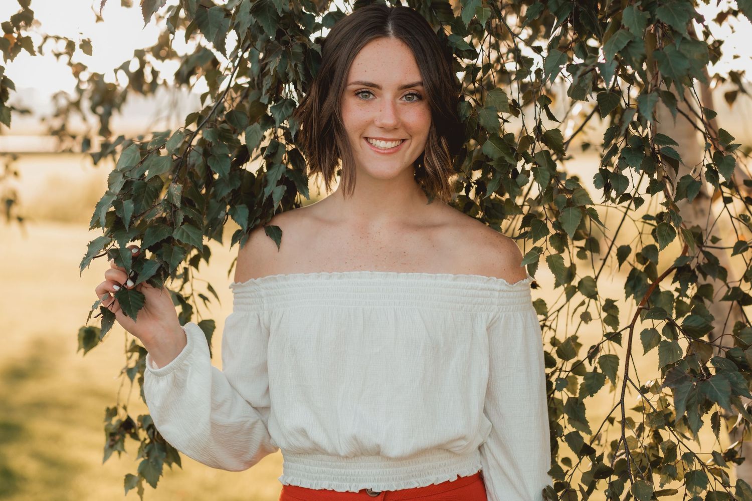 Smiling young woman in white off-shoulder top and red skirt standing among green leaves.