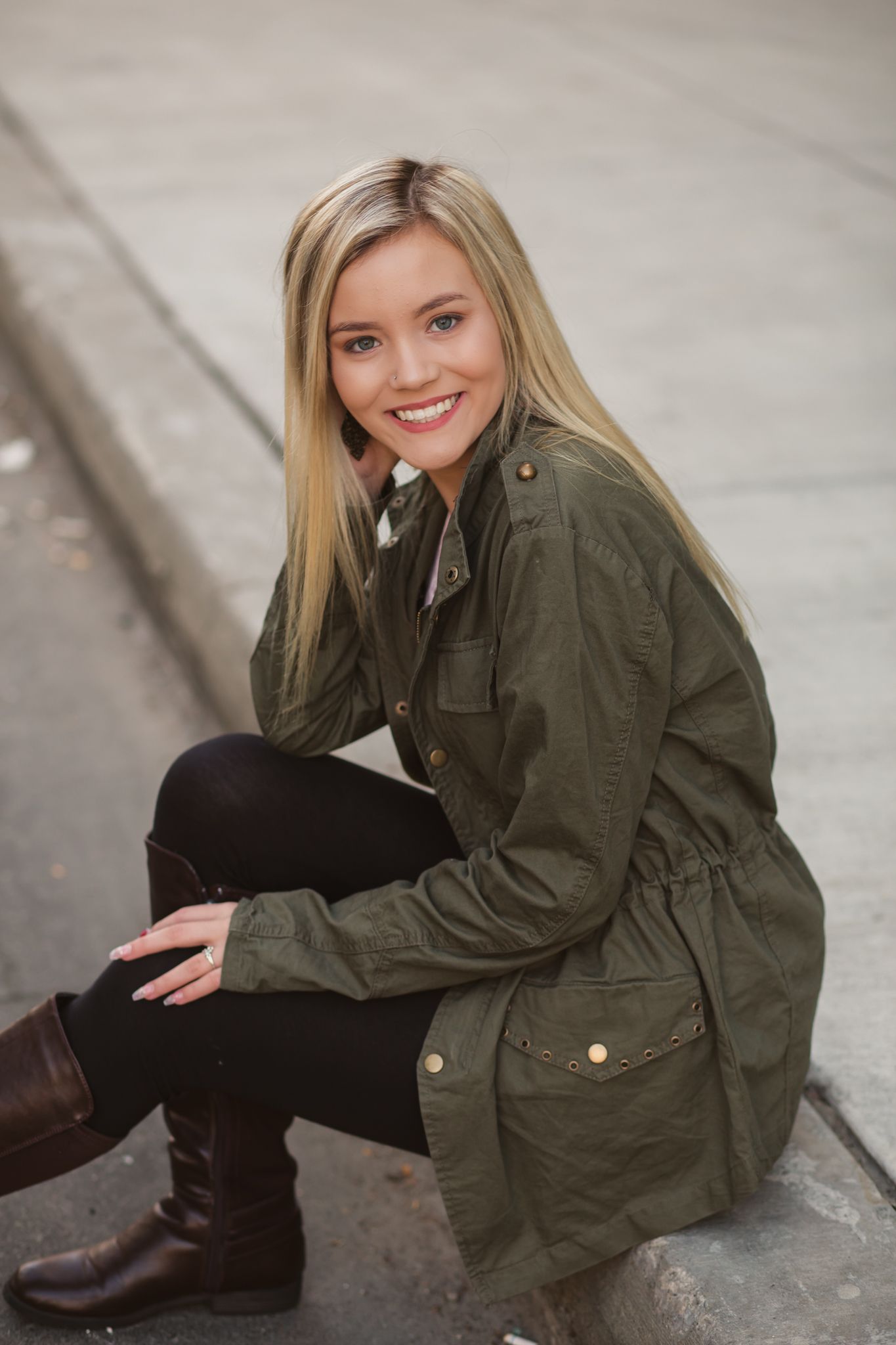 Smiling blonde woman in olive green jacket and brown boots sits on curb in urban setting.
