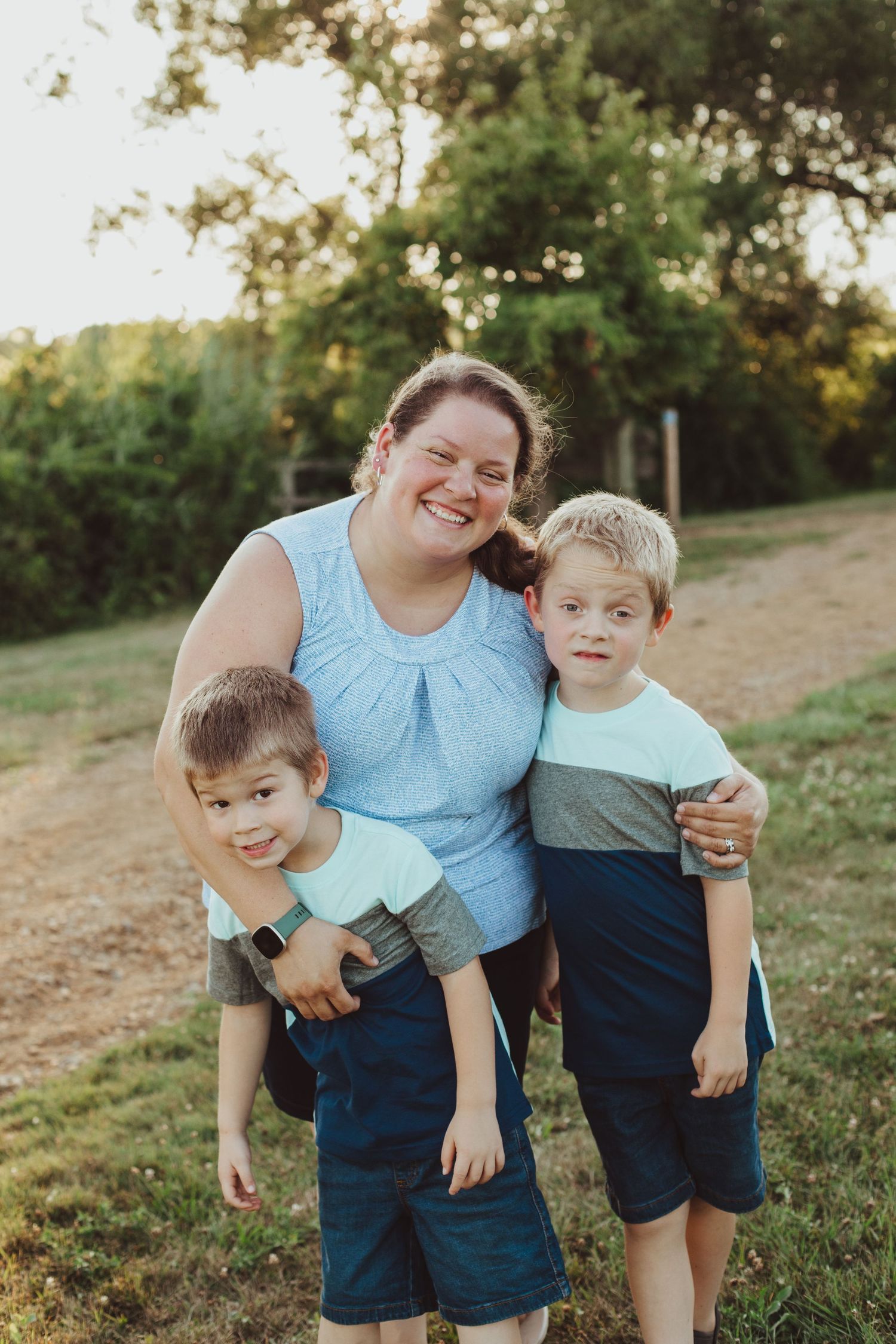 A smiling mother poses outdoors with her two young sons in a sunny green field at golden hour.