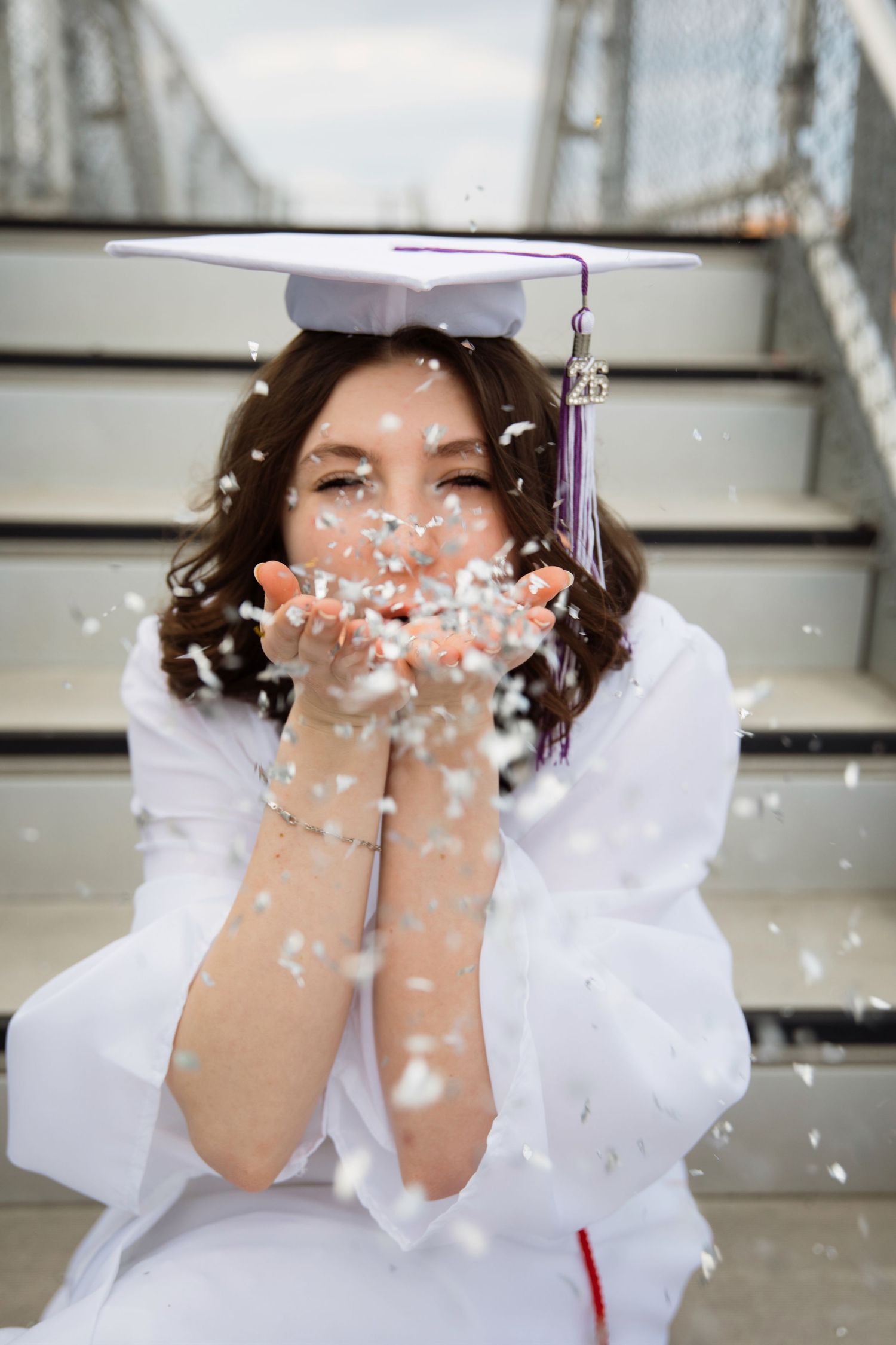 A young woman in white graduation gown and cap blows confetti from her hands while sitting on steps.