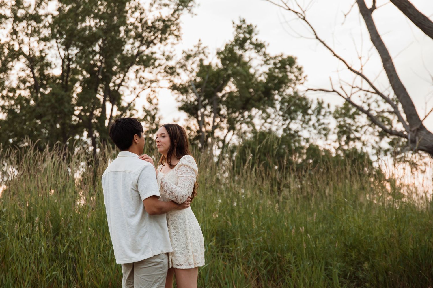 Couple embracing in tall grass field at sunset, sharing a romantic moment among lush green trees.