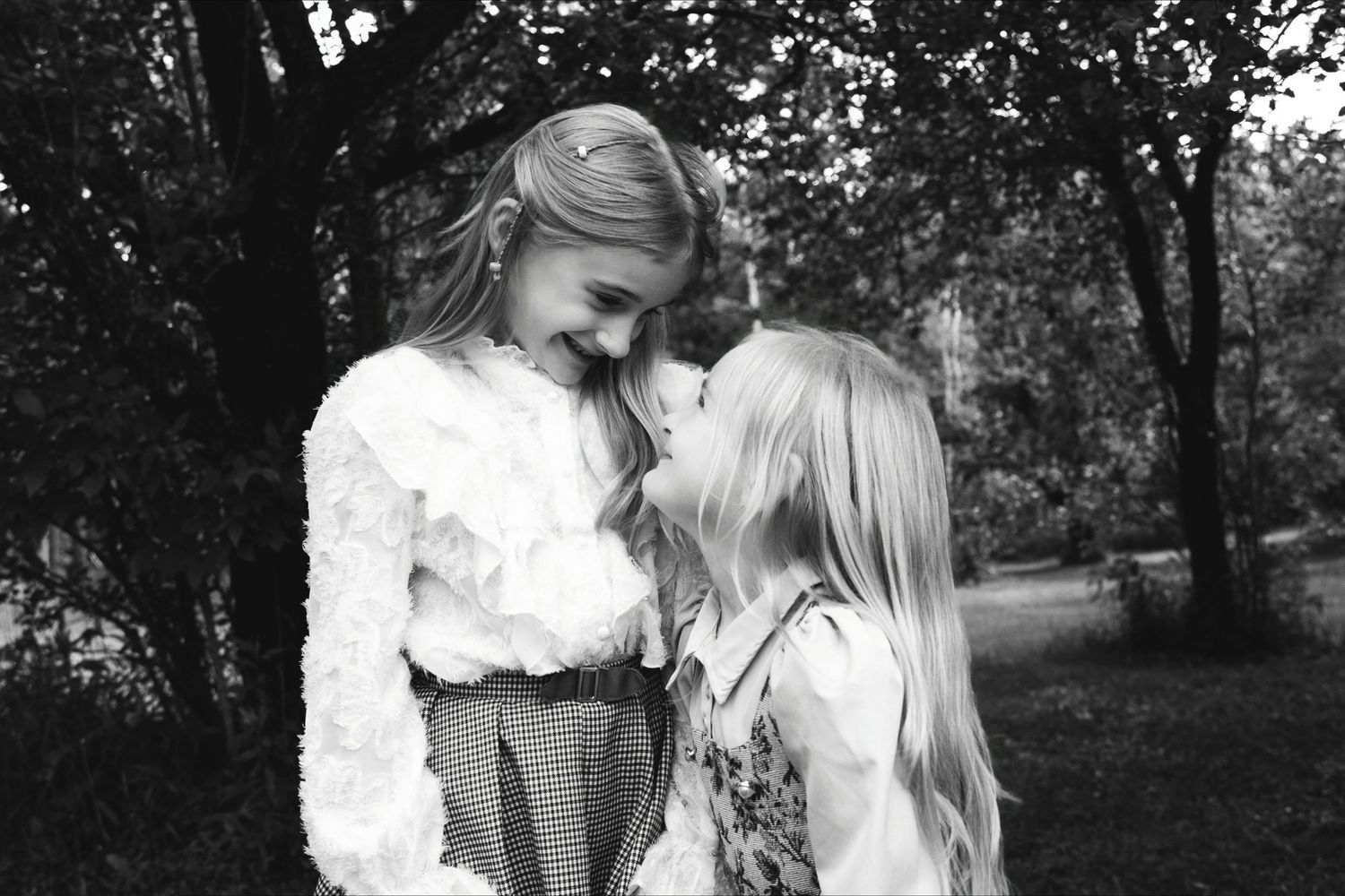 Two young girls share a tender moment in a black and white outdoor photo among trees.
