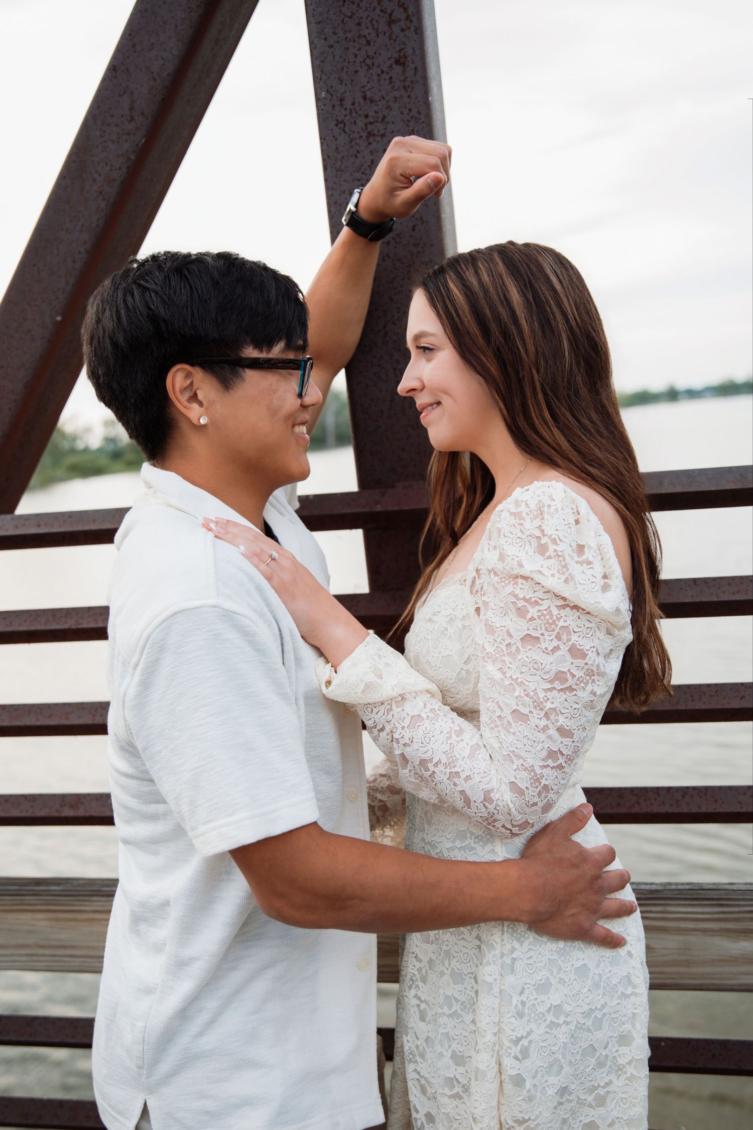 Couple embracing and smiling on a bridge near water, woman in lace dress, man in white shirt.