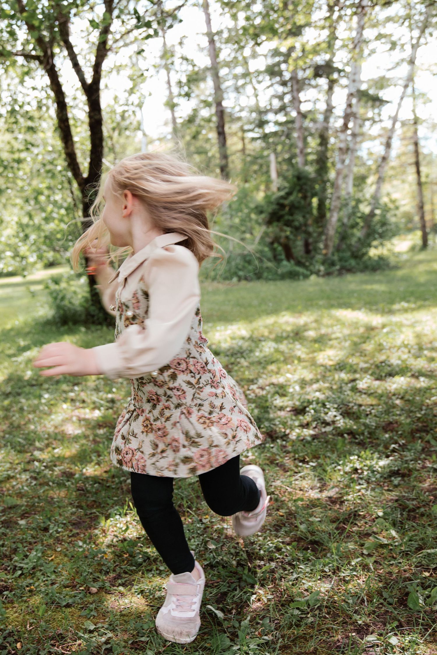 A young blonde girl in a floral dress and black leggings runs playfully through a lush green park.