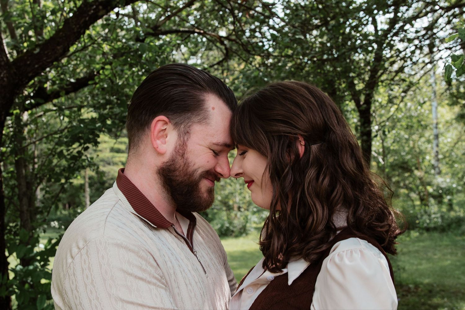 A romantic couple shares a tender moment touching foreheads outdoors surrounded by lush green trees.