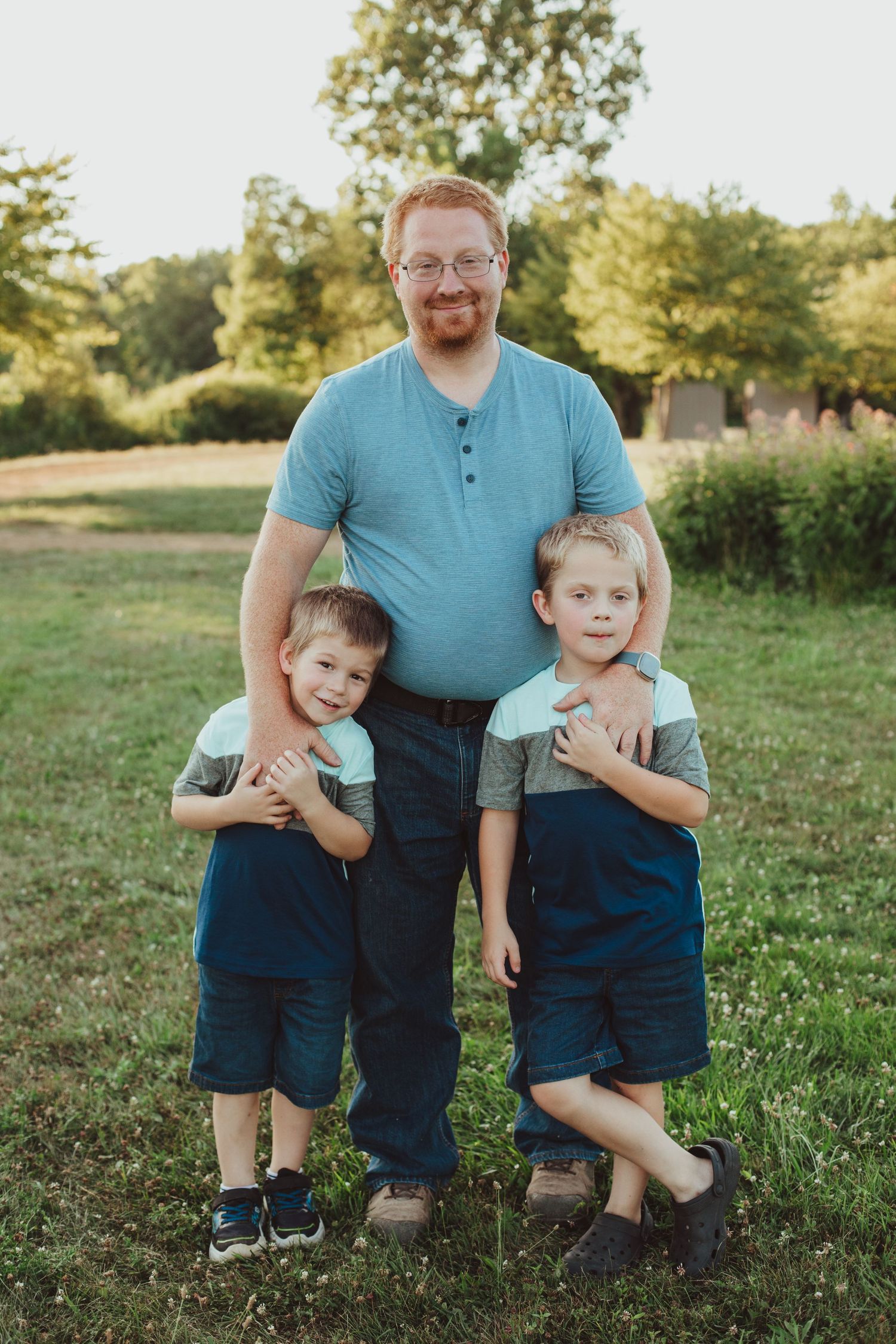 A red-haired father with glasses smiles while two young boys hug him in a sunny outdoor setting.