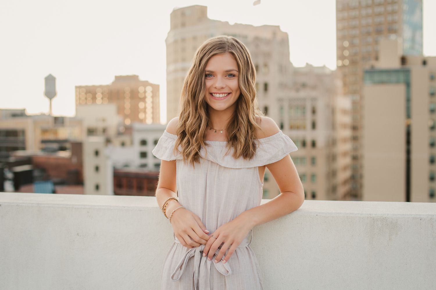 Smiling young woman in gray off-shoulder dress poses on rooftop with city skyline.