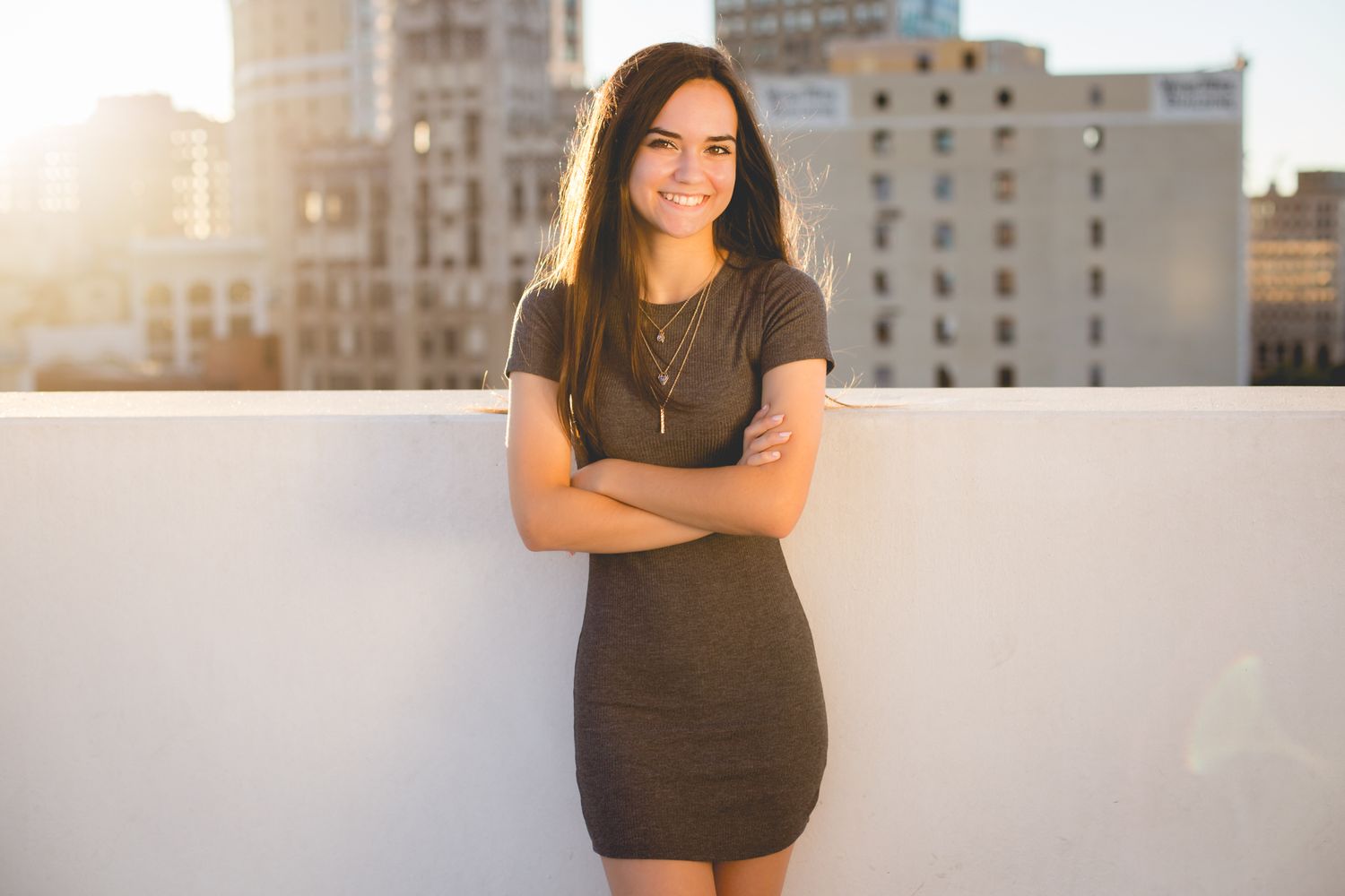 Smiling young woman in gray bodycon dress poses with arms crossed on sunny urban rooftop.