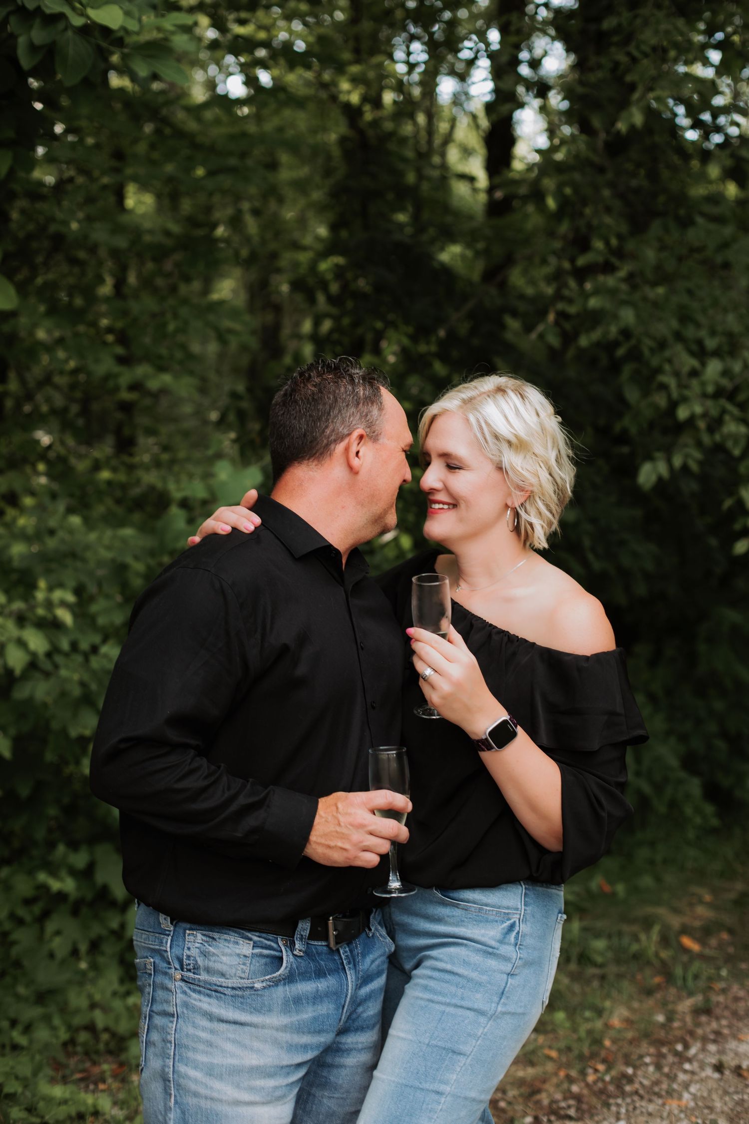 A romantic couple in black outfits toast champagne glasses while sharing an intimate moment outdoors.