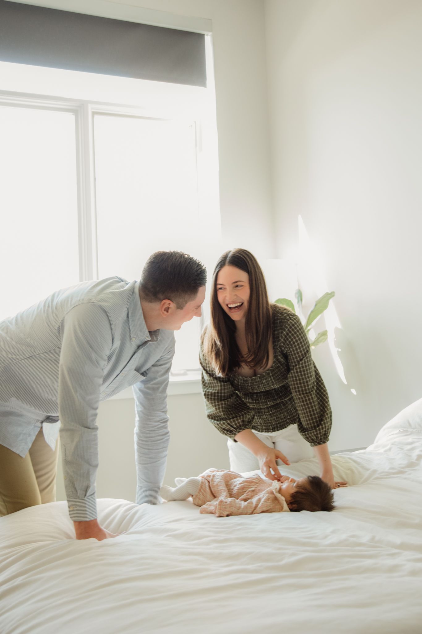 Laughing parents leaning over newborn baby lying on white bed in bright room.