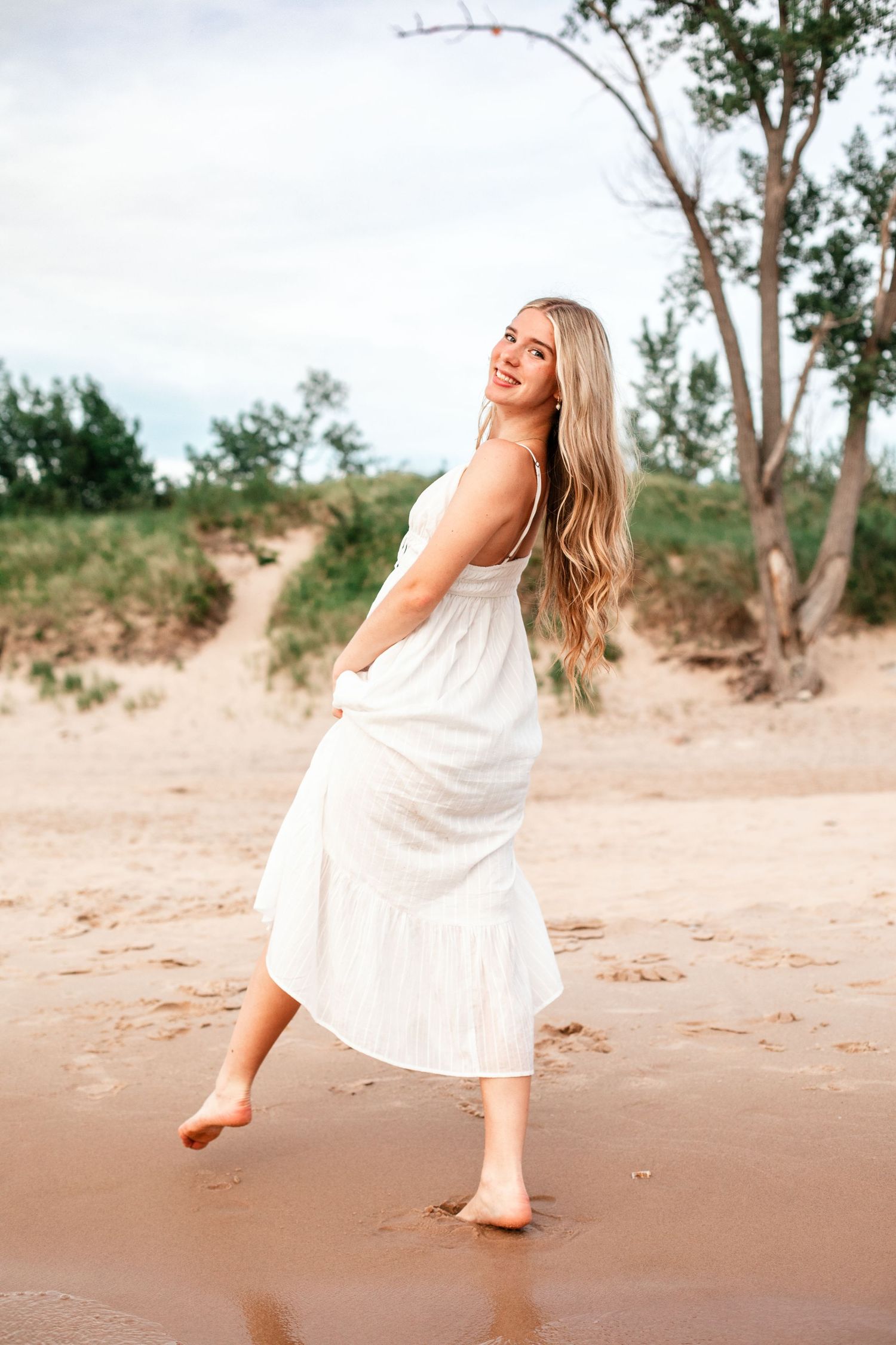 Young blonde woman in white sundress walking barefoot on sandy beach, smiling over shoulder.