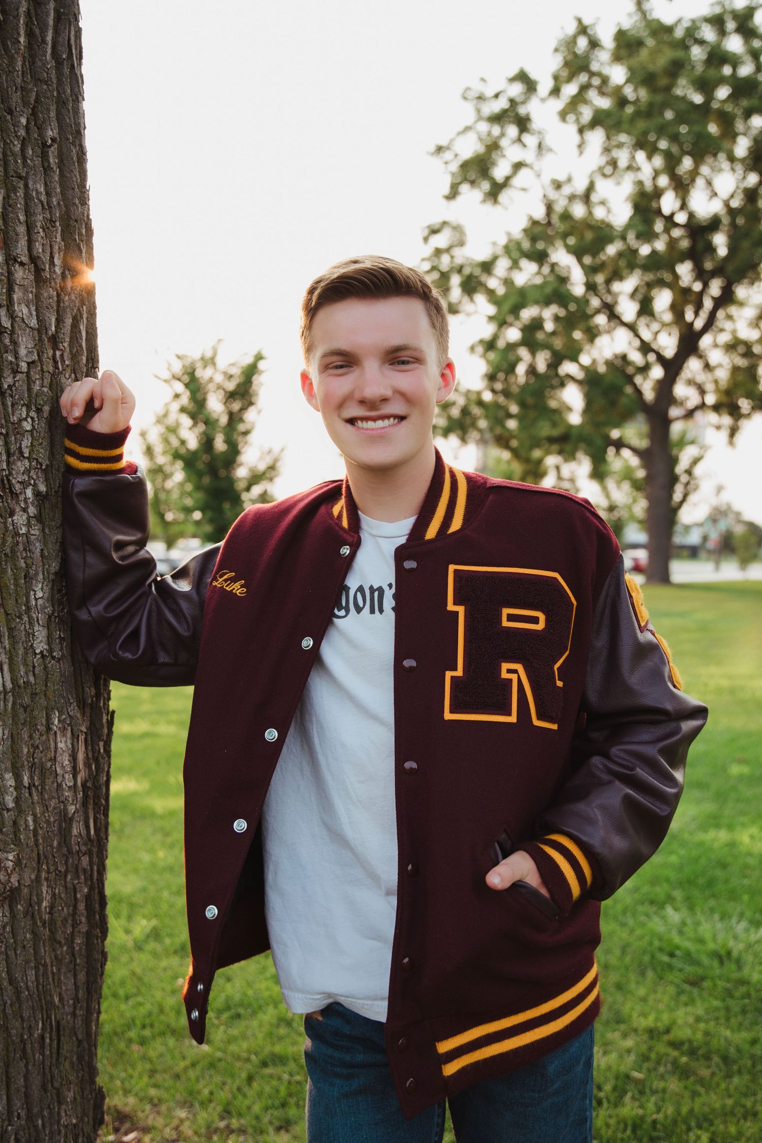 Smiling young man in maroon varsity jacket with R letter leaning against tree in park setting.