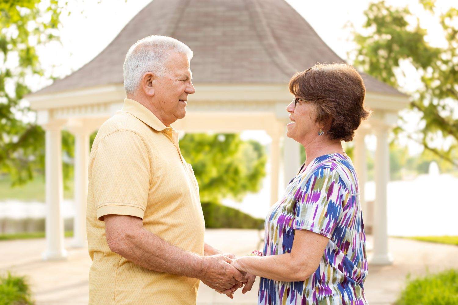 Elderly couple holding hands and facing each other in a park near a white gazebo on a sunny day.