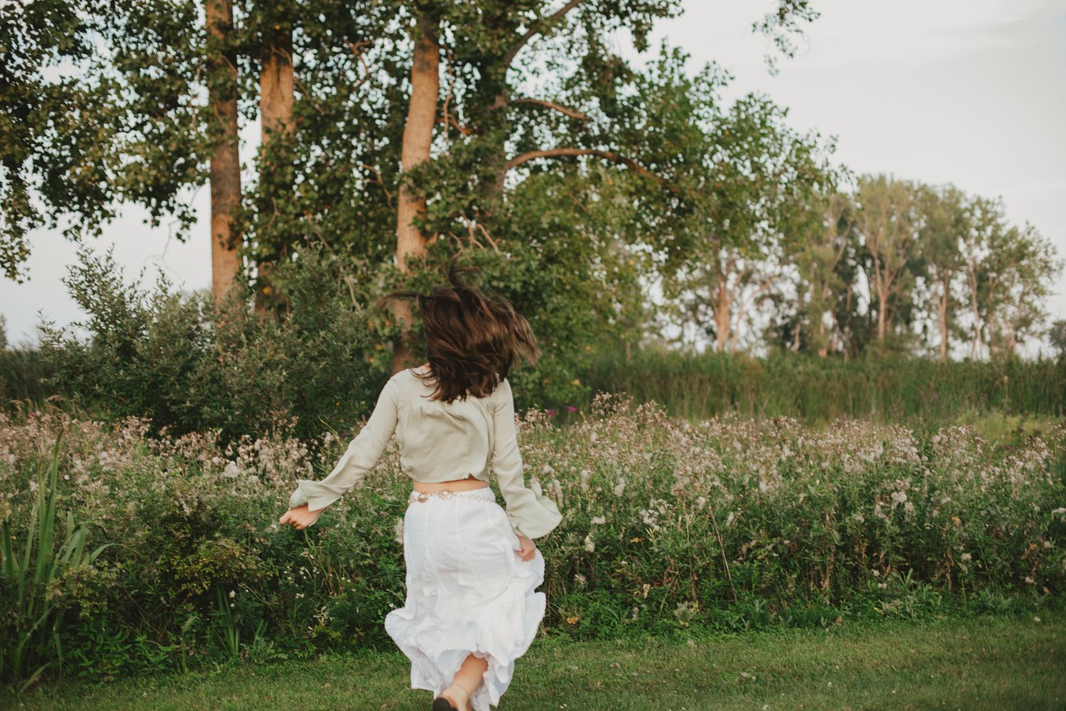A woman in a white skirt and green top twirls joyfully in a lush meadow surrounded by wildflowers and trees.