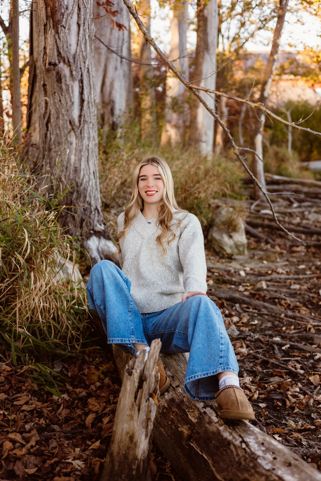 Smiling young woman in cream sweater and jeans sitting on a log in autumn forest setting.