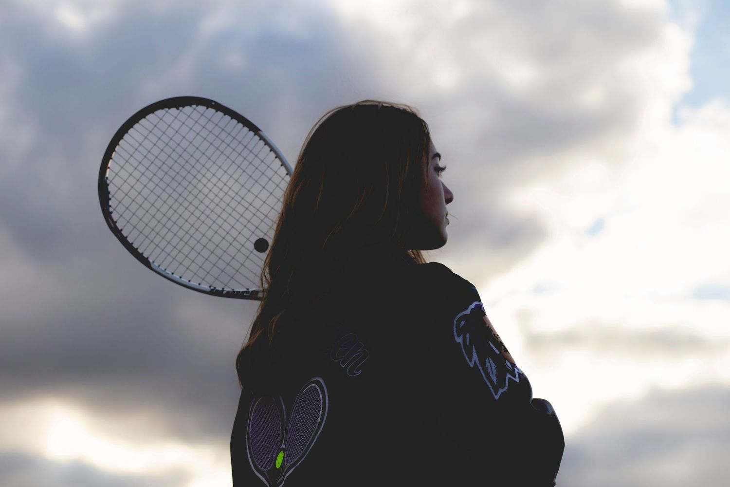 A young woman holds a tennis racket over her shoulder against a dramatic cloudy sky backdrop.
