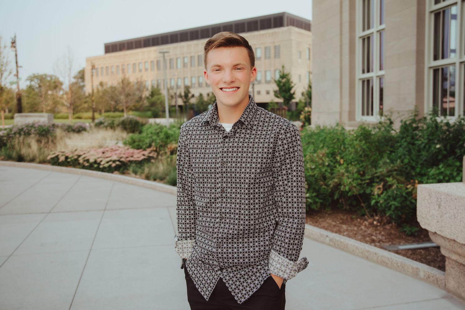 Smiling young man in patterned shirt posing on campus walkway with brick building in background.
