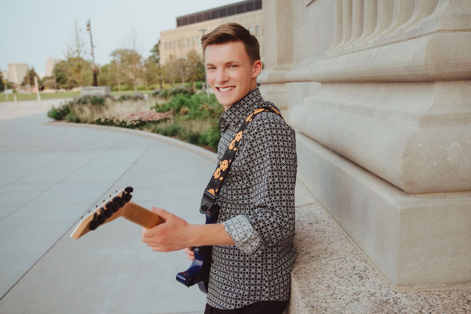 Smiling young man holding blue electric guitar leaning against stone building.