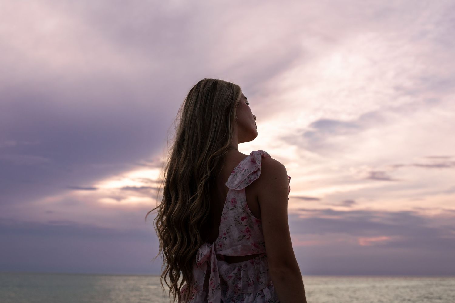 Young woman with long wavy hair gazes at a stunning purple sunset over the ocean in a floral dress.