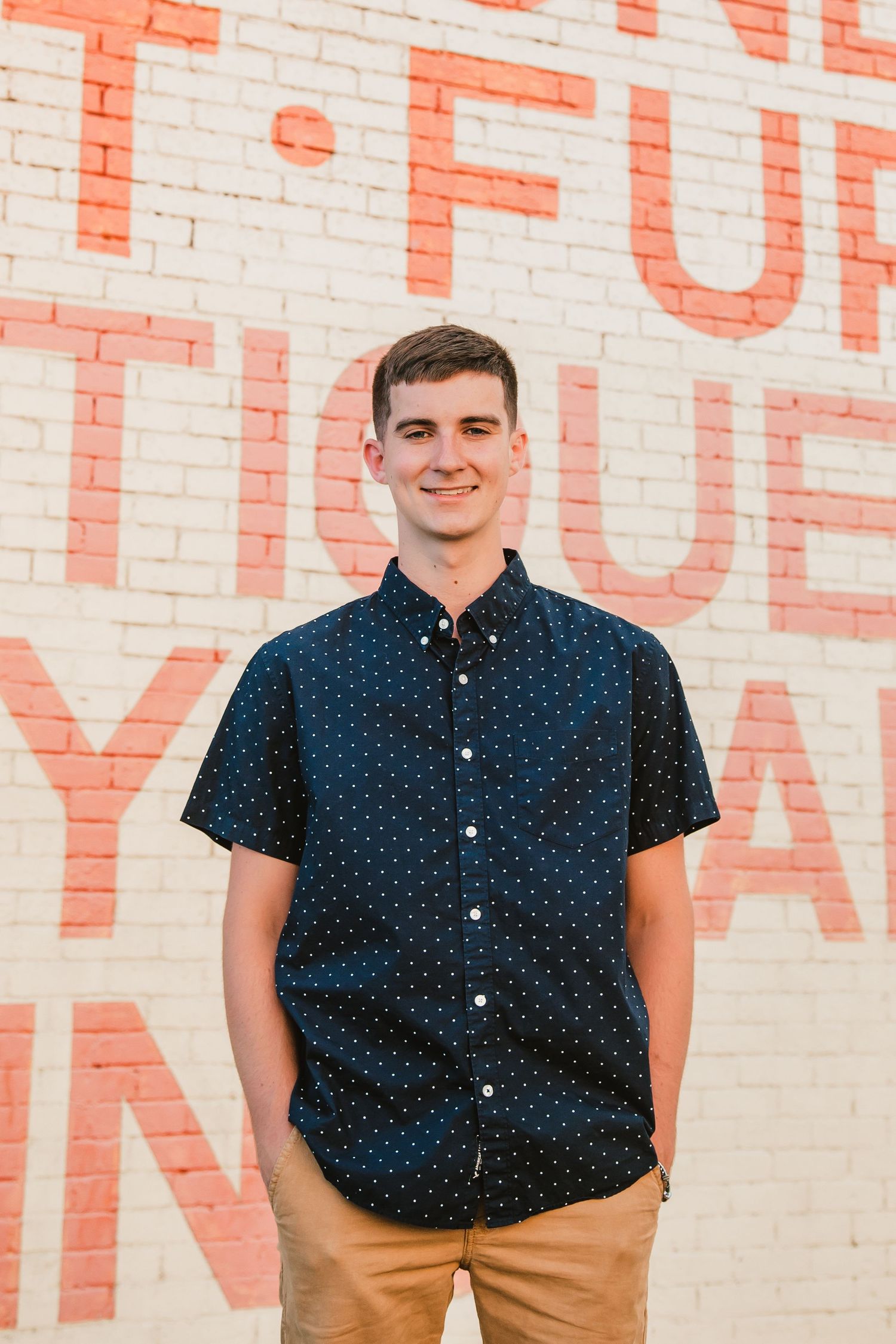 Young man smiling in navy polka dot shirt and khaki pants standing against a white brick wall with red painted lettering.
