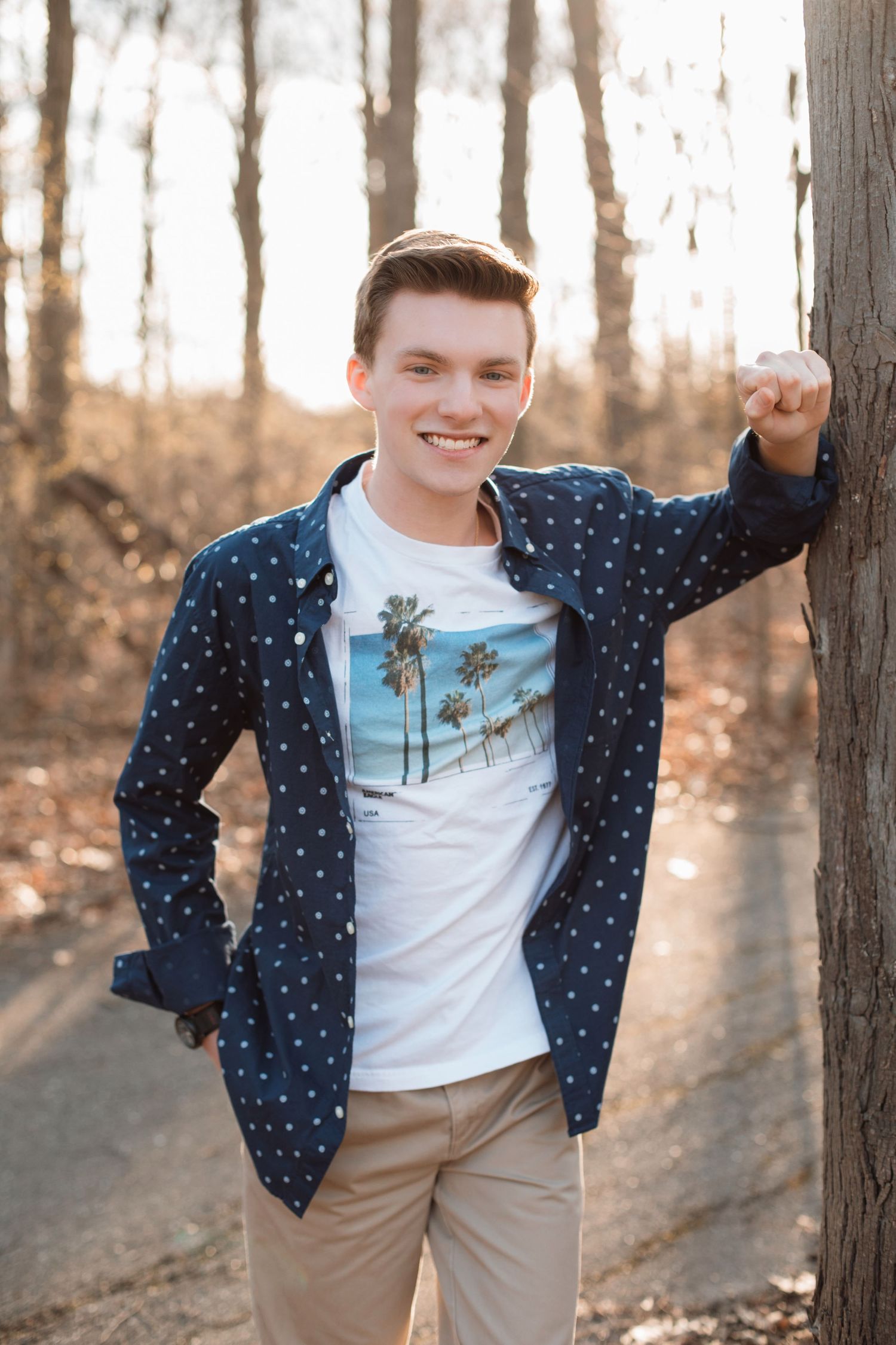 Smiling young man in a navy polka dot shirt and palm tree tee leans against a tree in a sunlit forest.