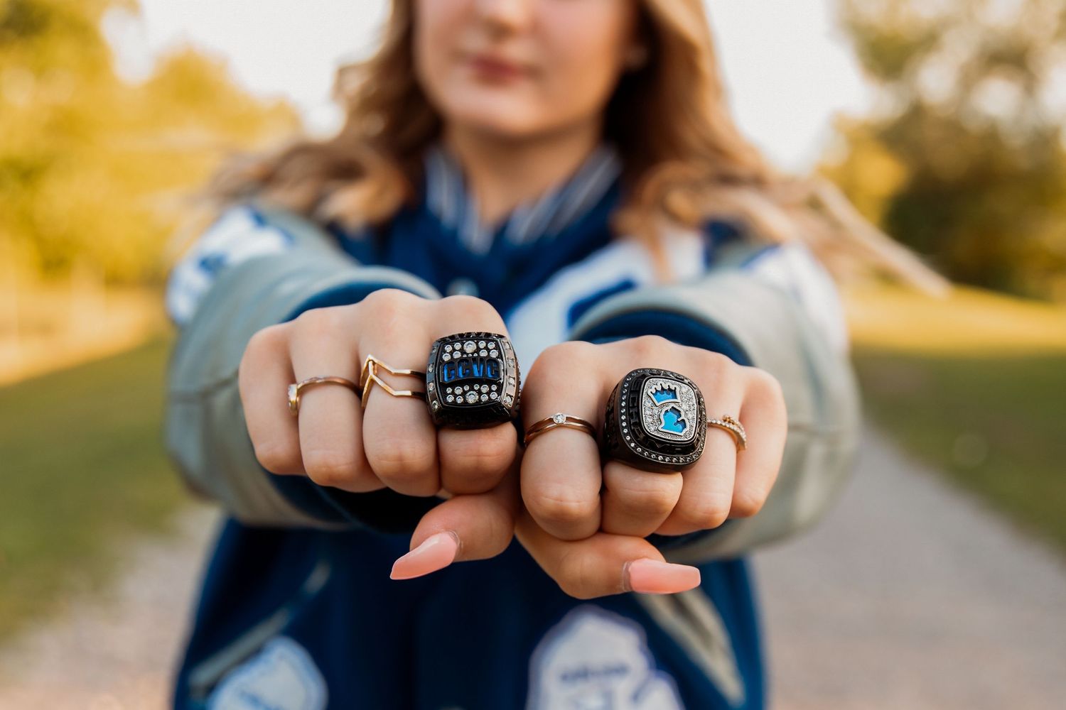 Woman proudly displaying two large championship rings on her fists outdoors, wearing a blue jacket.