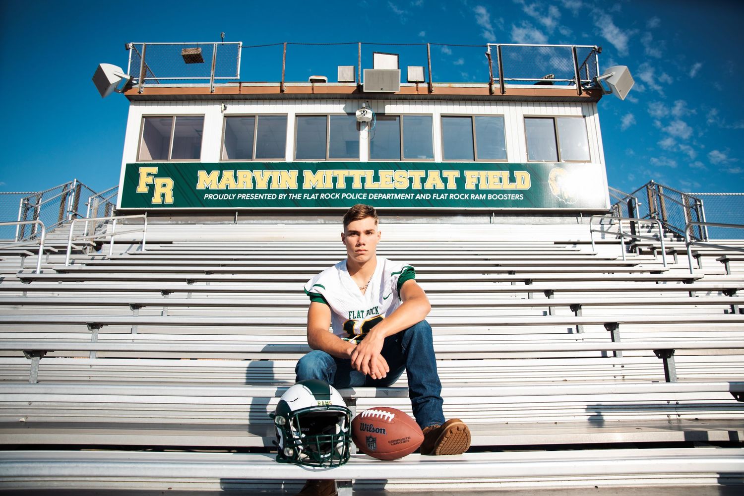 Young football player poses on bleachers at Marvin Mittlestat Field, holding a helmet and football under blue skies.