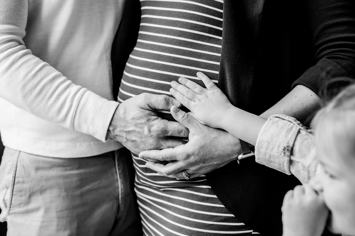 Black and white close-up of family hands resting on a pregnant belly, symbolizing love and new life.