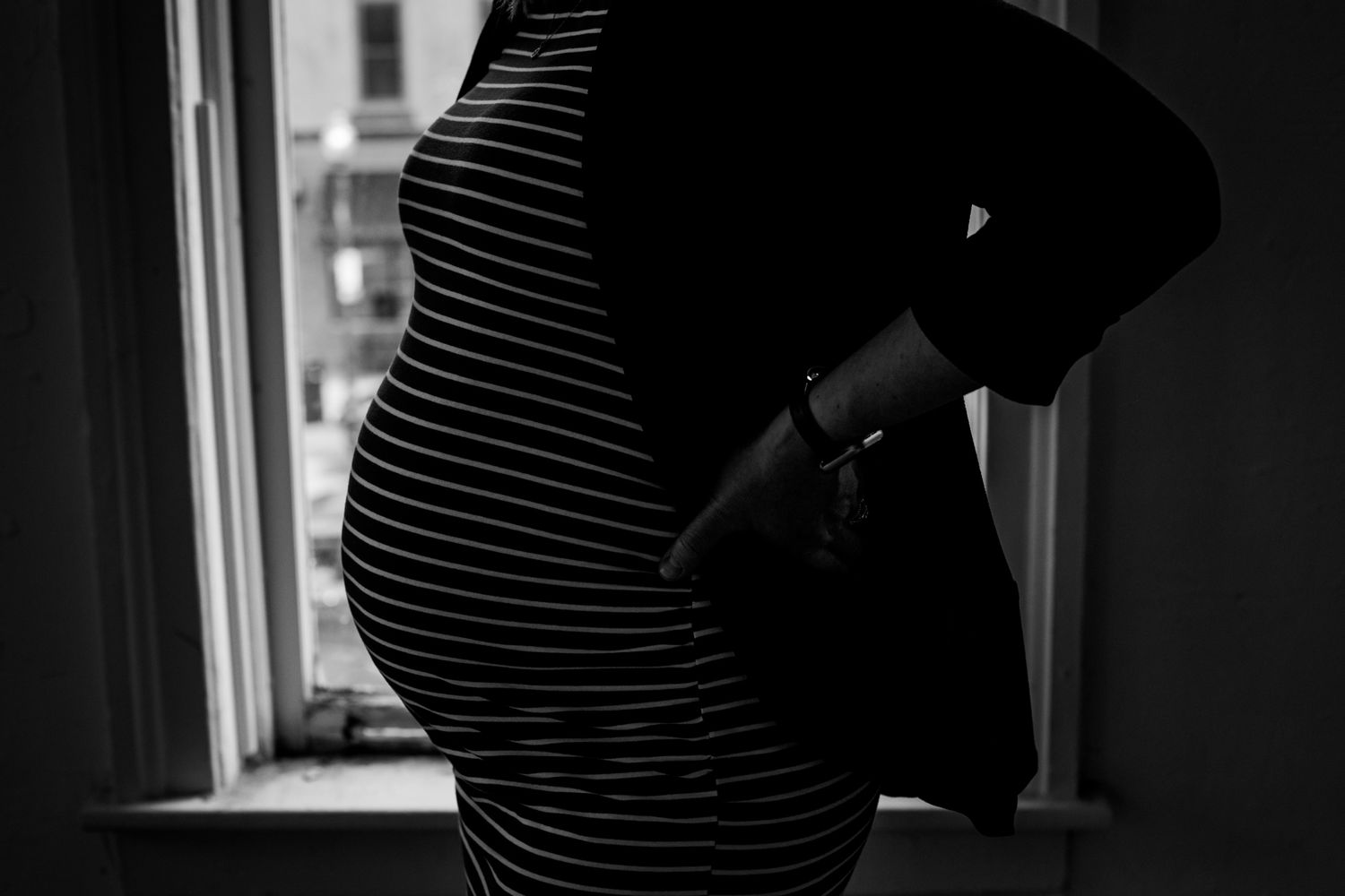 Close-up black and white photo of pregnant woman in striped dress cradling her baby bump by window.