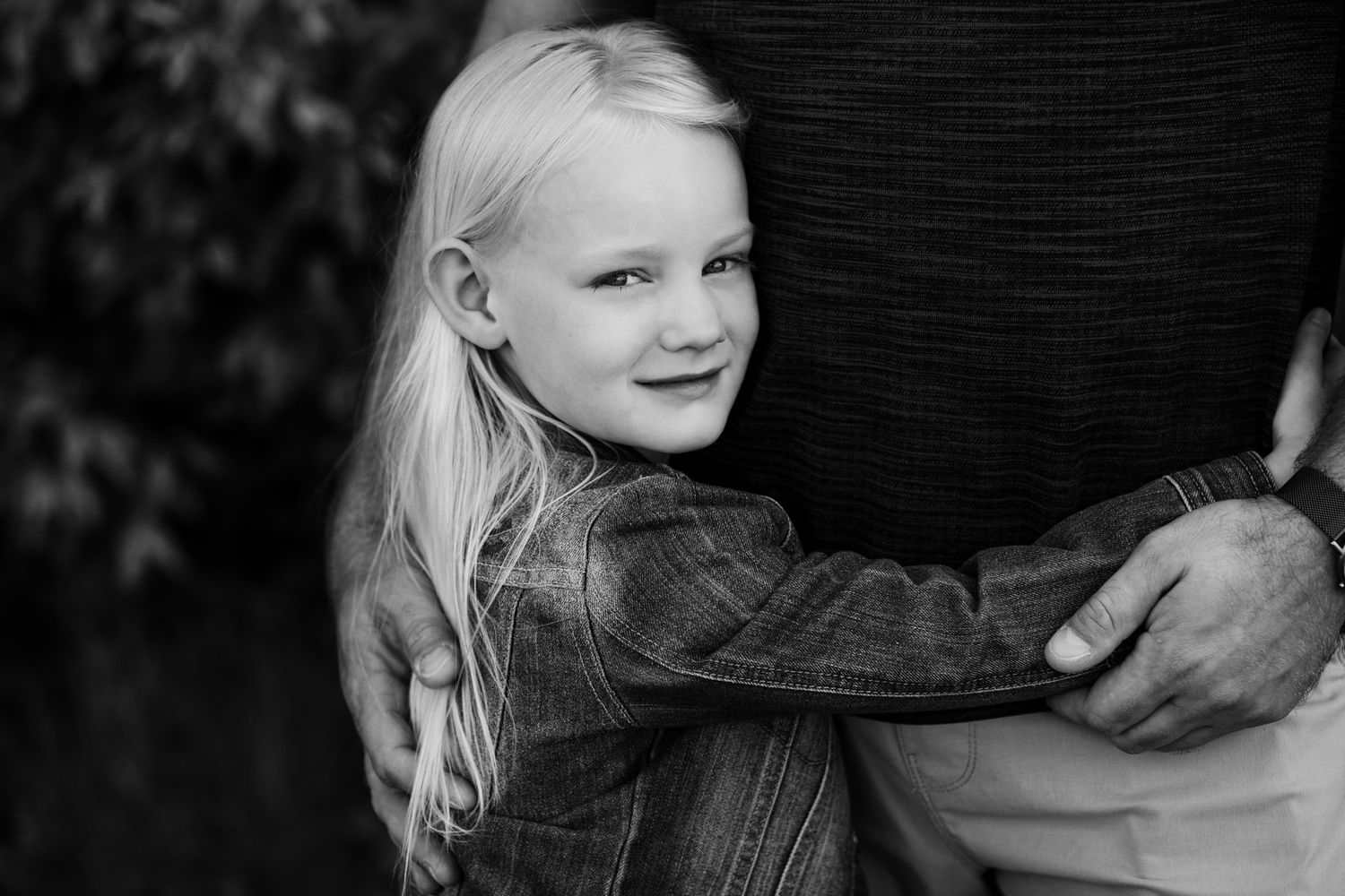 Black and white photo of a smiling blonde girl hugging an adult outdoors.