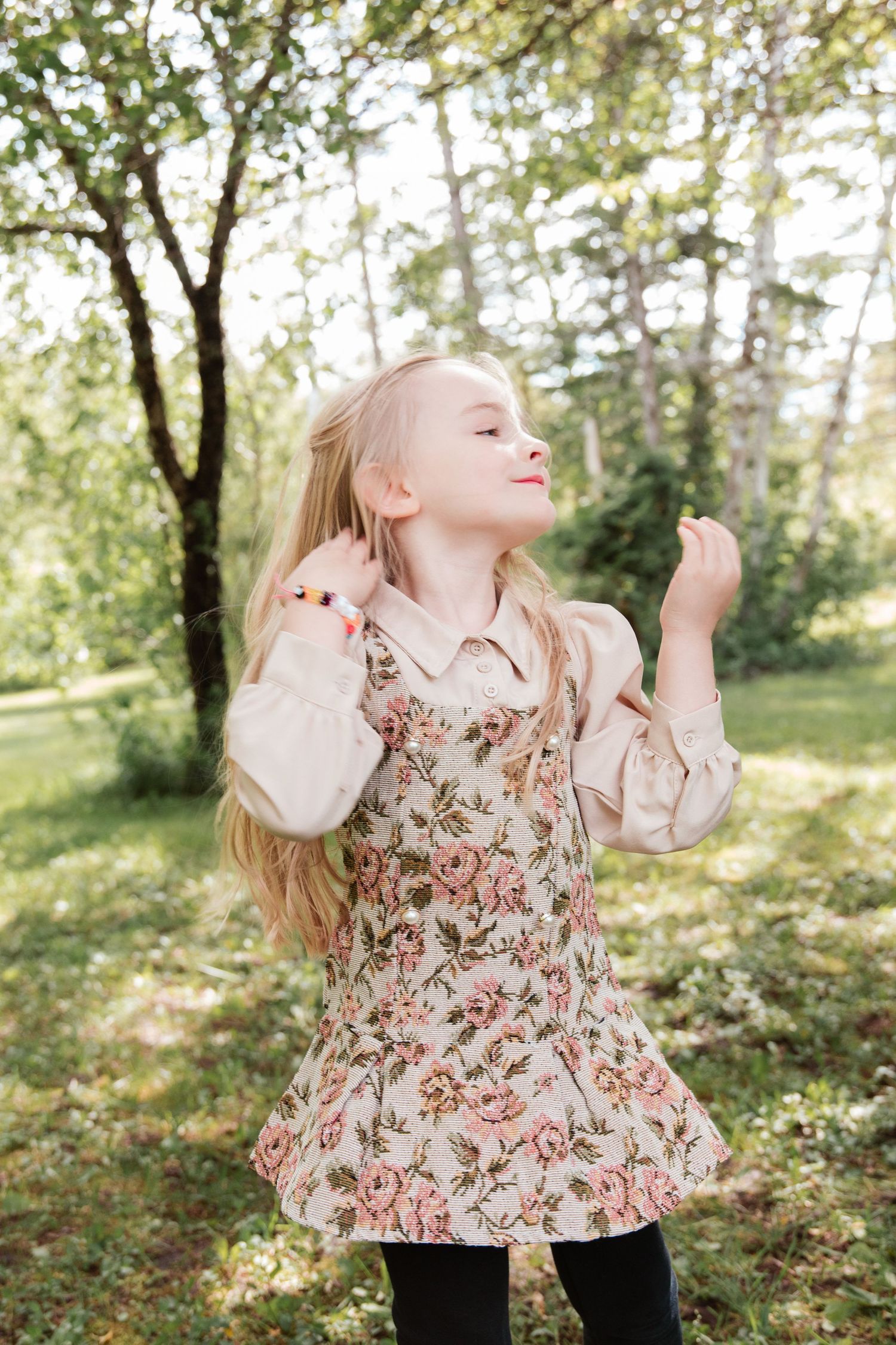 Young blonde girl in floral dress dancing outdoors in a sunny green park, smiling with eyes closed.