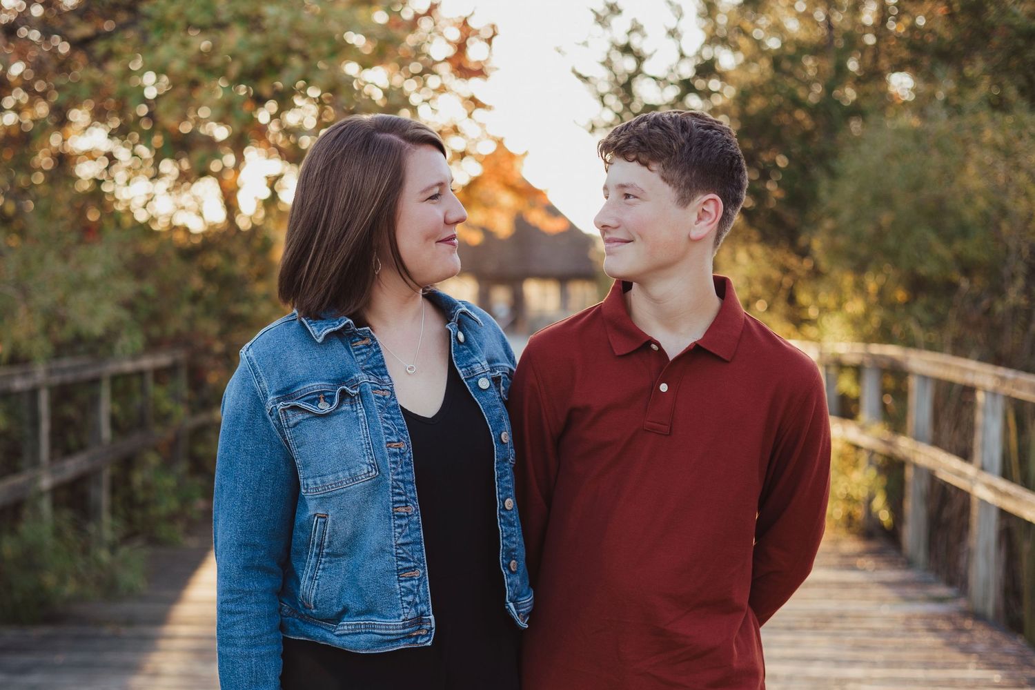 A young woman in a denim jacket and a teen boy in a red polo smile at each other on a wooden bridge outdoors.