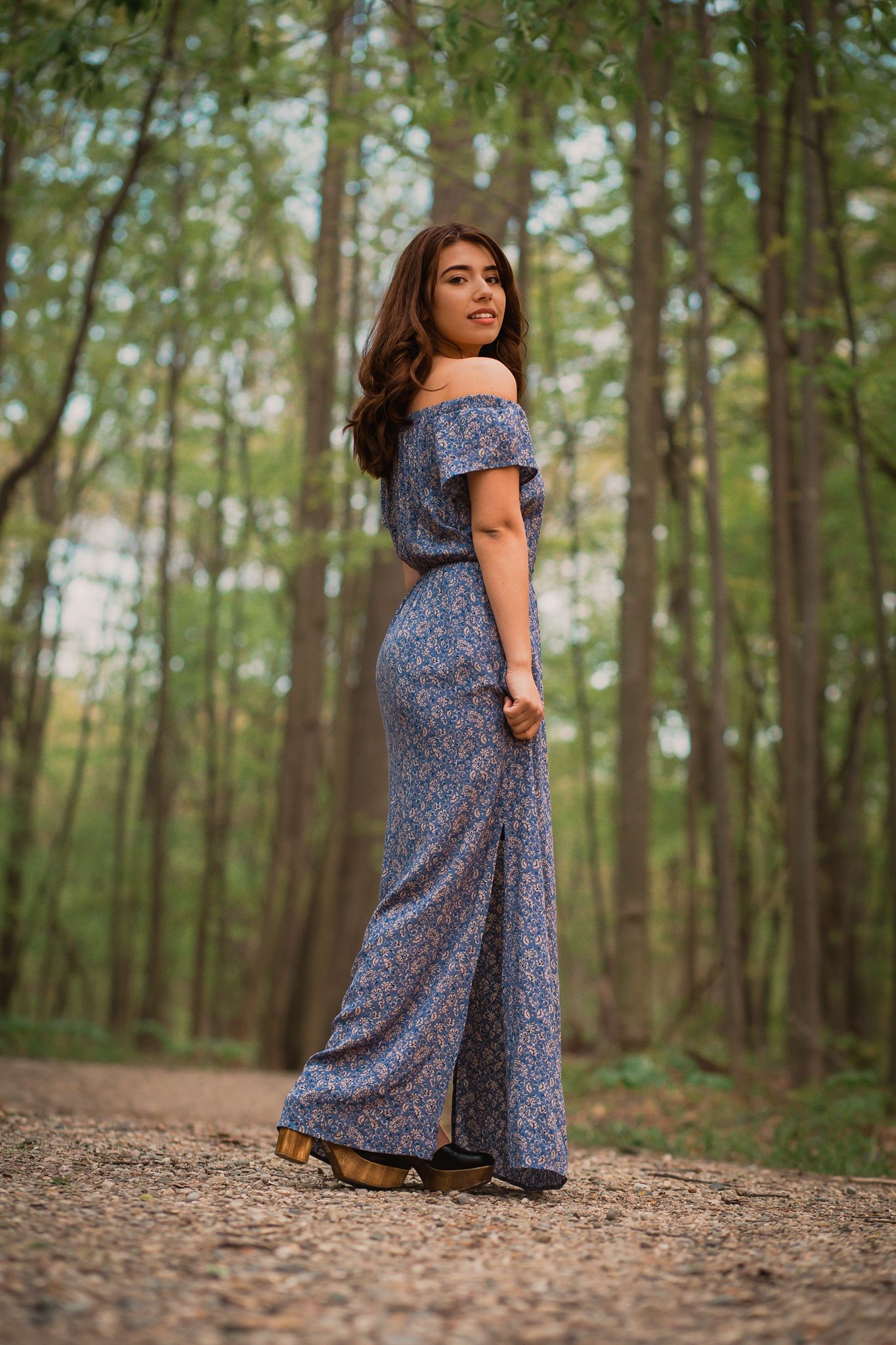 Brunette woman in blue floral off-shoulder maxi dress standing on forest path among tall trees.